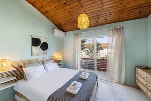 A bedroom with a bed, bedside tables, a hanging light fixture, a window with curtains, and a view of the outside. - Laranca Oneiro Seaside Villa (Pervolia)