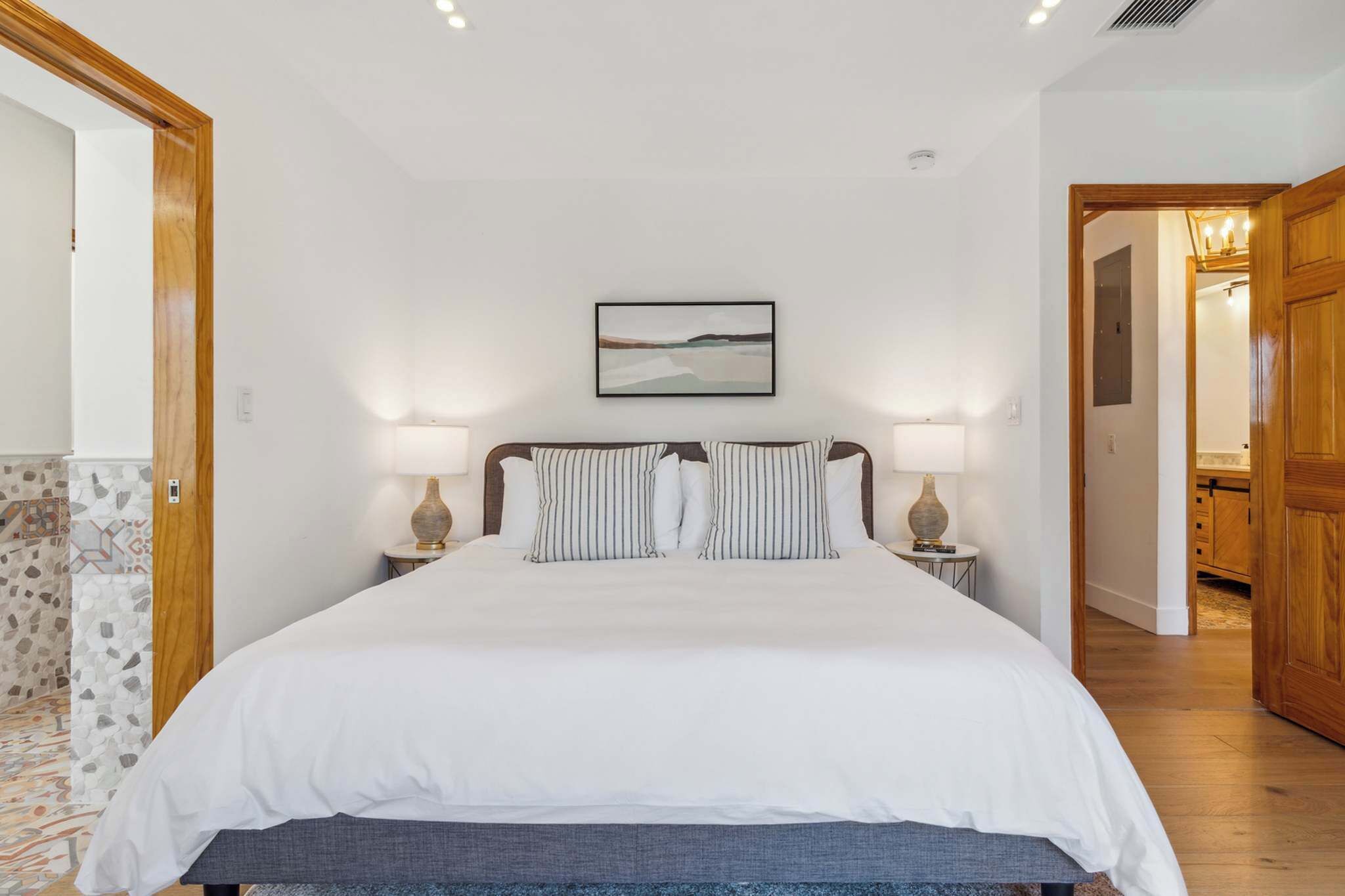 A bedroom with a large bed, two bedside tables with lamps, a framed picture on the wall, and a wooden door leading to another room.