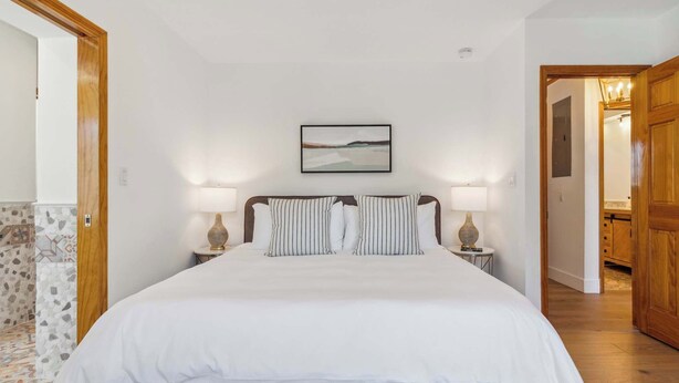 Une chambre avec un grand lit, deux tables de chevet avec des lampes, un tableau encadré au mur et une porte en bois donnant sur une autre pièce.