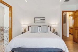 A bedroom with a large bed, two bedside tables with lamps, a framed picture on the wall, and a wooden door leading to another room.