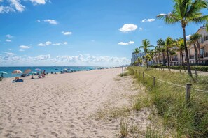 A sandy beach with umbrellas and people, palm trees, and buildings in the background.