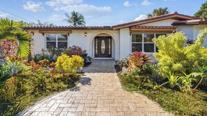 Une maison à deux étages avec une allée pavée menant à la porte d’entrée, entourée de diverses plantes tropicales et surplombant un ciel bleu azur.