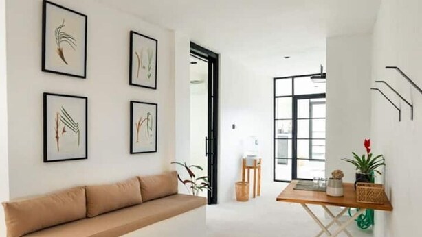A modern interior with a beige sofa, framed botanical prints, a wooden side table, and a view through a doorway.