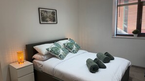 Una cama bien hecha con ropa blanca y almohadas con estampado de hojas verdes, una lámpara de noche y toallas enrolladas sobre la cama.