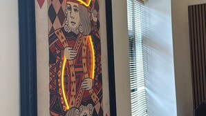 Ein gerahmtes Spielkartemotiv mit Neonumrandung, ein Fenster mit Jalousien und eine Pflanze auf einem Fensterbrett.