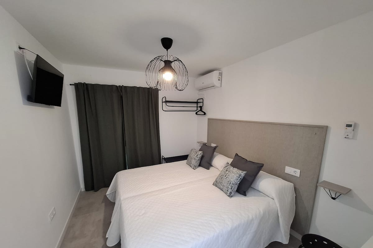 A neatly arranged bedroom with a bed, a hanging light fixture, an air conditioning unit, and a television mounted on the wall.