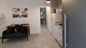 Un salon moderne avec un canapé, un coin cuisine équipé d’un réfrigérateur, et un couloir menant à une salle de bain.