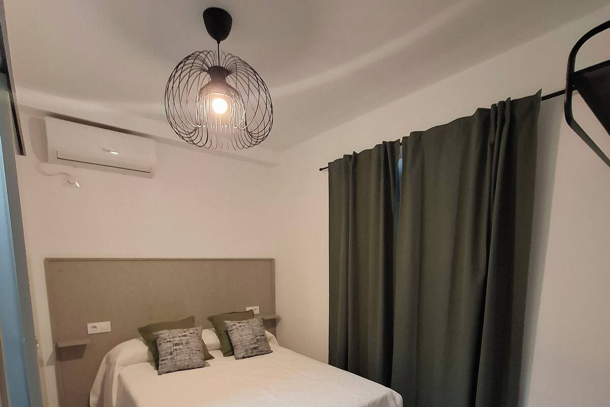 A bedroom with a bed, a desk, a chair, a hanging light fixture, and a window with curtains.