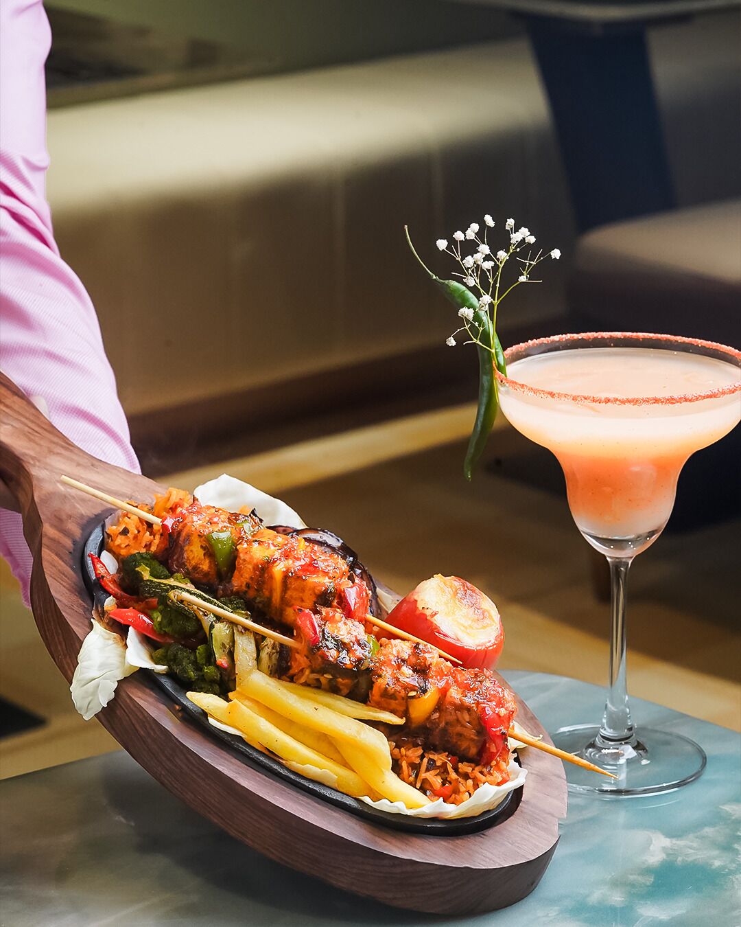 Een dienblad met gegrilde satés en groenten, een roze cocktail en een klein bloemetje op een tafel.