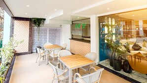 Un comedor moderno con mesas de madera y sillas de mimbre, un papel tapiz decorativo y vista a la calle a través de un ventanal.