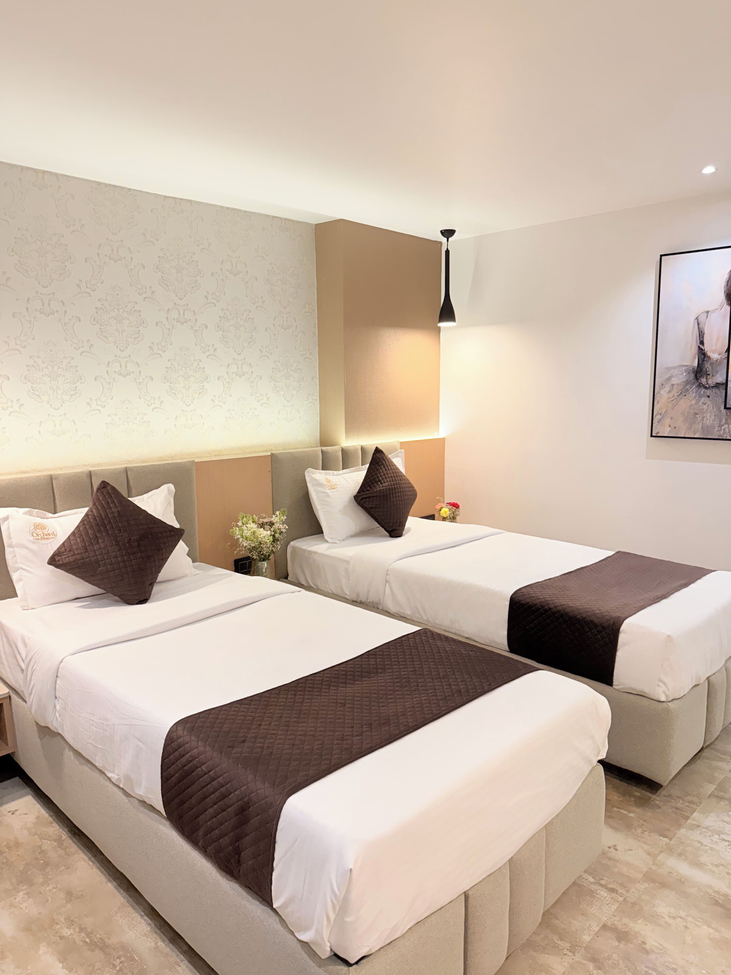 Une chambre d’hôtel avec deux lits, chacun recouvert d’une courtepointe blanche et marron, doté d’une tête de lit et agrémenté d’une table de chevet sur laquelle se trouve un vase de fleurs.