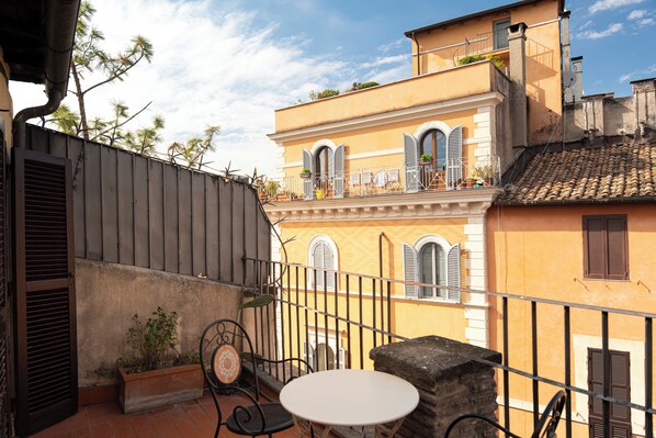 A balcony with a table and chairs, overlooking a building with a tiled roof and a yellow facade.