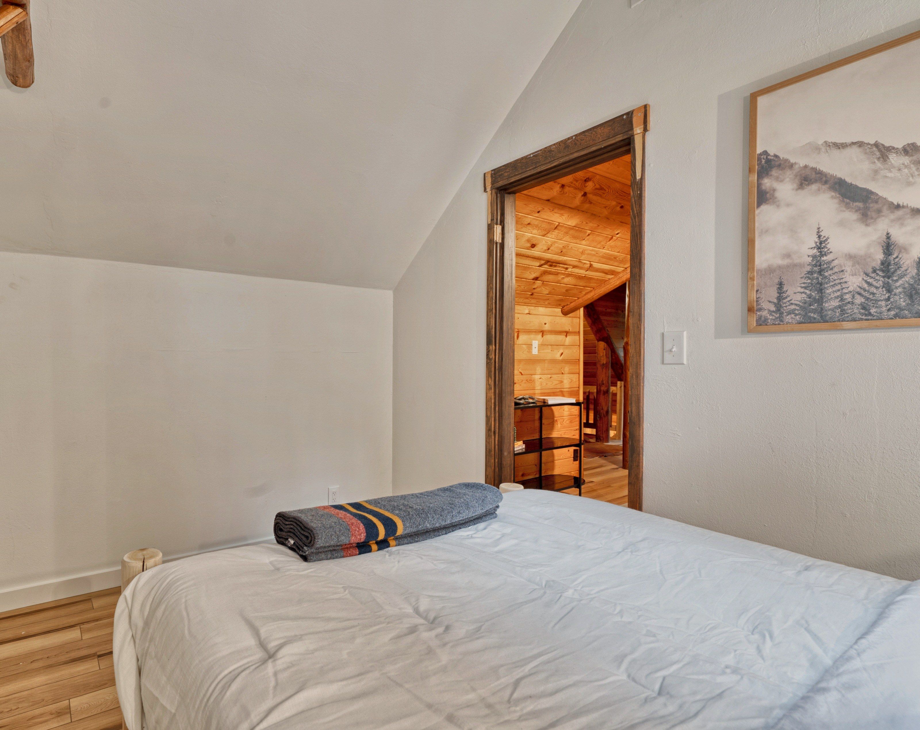 A bedroom with a bed, a wooden door leading to a wooden room, and a mountain landscape painting on the wall.