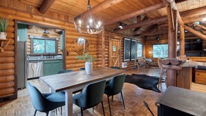 Interieur van een blokhut met een houten eettafel, stoelen, een kroonluchter, een keukenhoek en een groot raam.