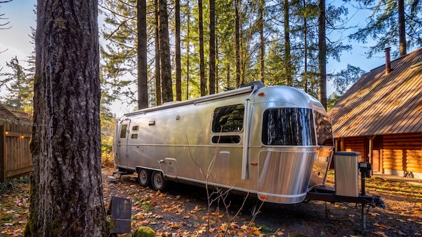 Un véhicule récréatif argenté est garé dans une zone boisée, avec une cabane en bois en arrière-plan.