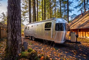 A silver recreational vehicle parked in a forested area with a wooden cabin in the background. - Log Cabin + Airstream | Hot Tub | Near Mt. Hood (Mount Hood Village)
