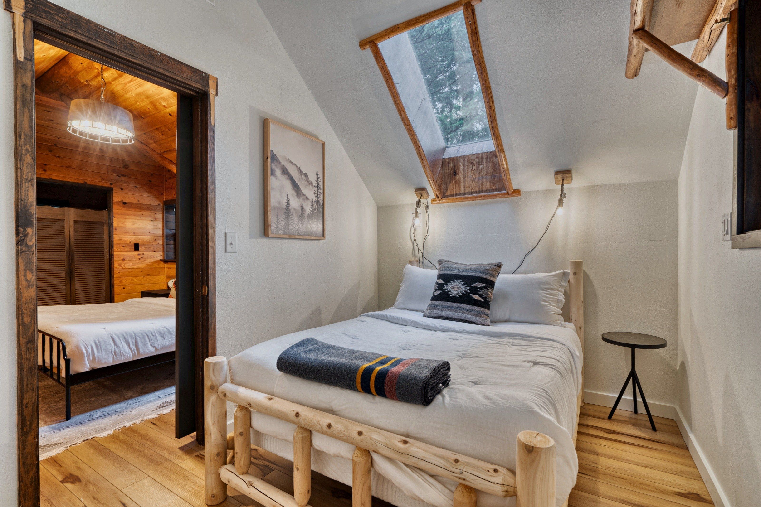 Een slaapkamer met een houten bedframe, een klein nachtkastje, een wandlamp en een raam met uitzicht op bomen.