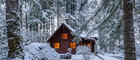 Een blokhut omringd door met sneeuw bedekte bomen, met een houten terras en een picknicktafel op de voorgrond.