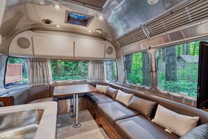 A modern RV interior with a dining area, seating, and large windows offering a view of the forest.