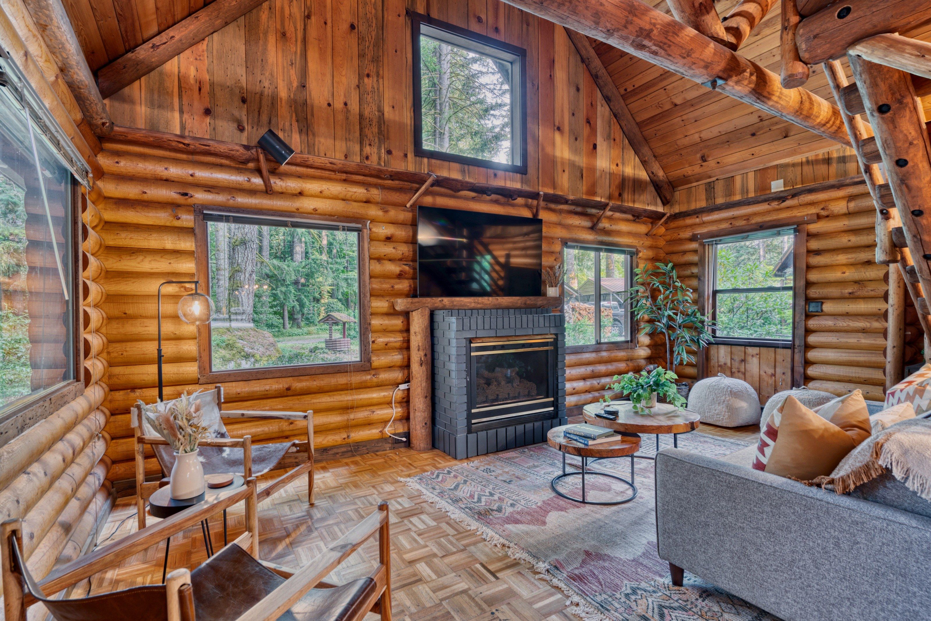 A log cabin interior with a fireplace, seating area, and large windows.