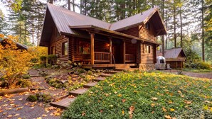 Une cabane en rondins dotée d’un porche et d’un avant-toit, entourée d’une forêt et d’un jardin aux feuilles d’automne.