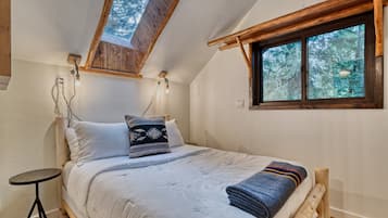 A bedroom with a sloped ceiling, a skylight, a bed with pillows, a bedside table, and a window with a view of trees.