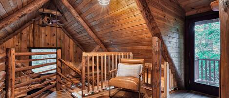 A wooden interior with a sloped ceiling, a wooden chair, a hanging light fixture, and a view through a window.