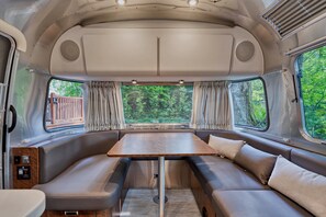 Wohnmobil-Innenausstattung mit Esstisch, gepolsterten Sitzen und großen Fenstern, die einen Blick ins Grüne bieten.