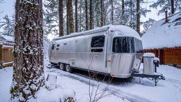 A stationary RV parked in a snowy forested area with a wooden fence and a wooden building in the background.