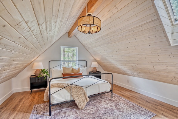 A bedroom with a sloped ceiling, a bed with a metal frame, two bedside tables with lamps, a window with a view of trees, and a patterned rug on the floor.
