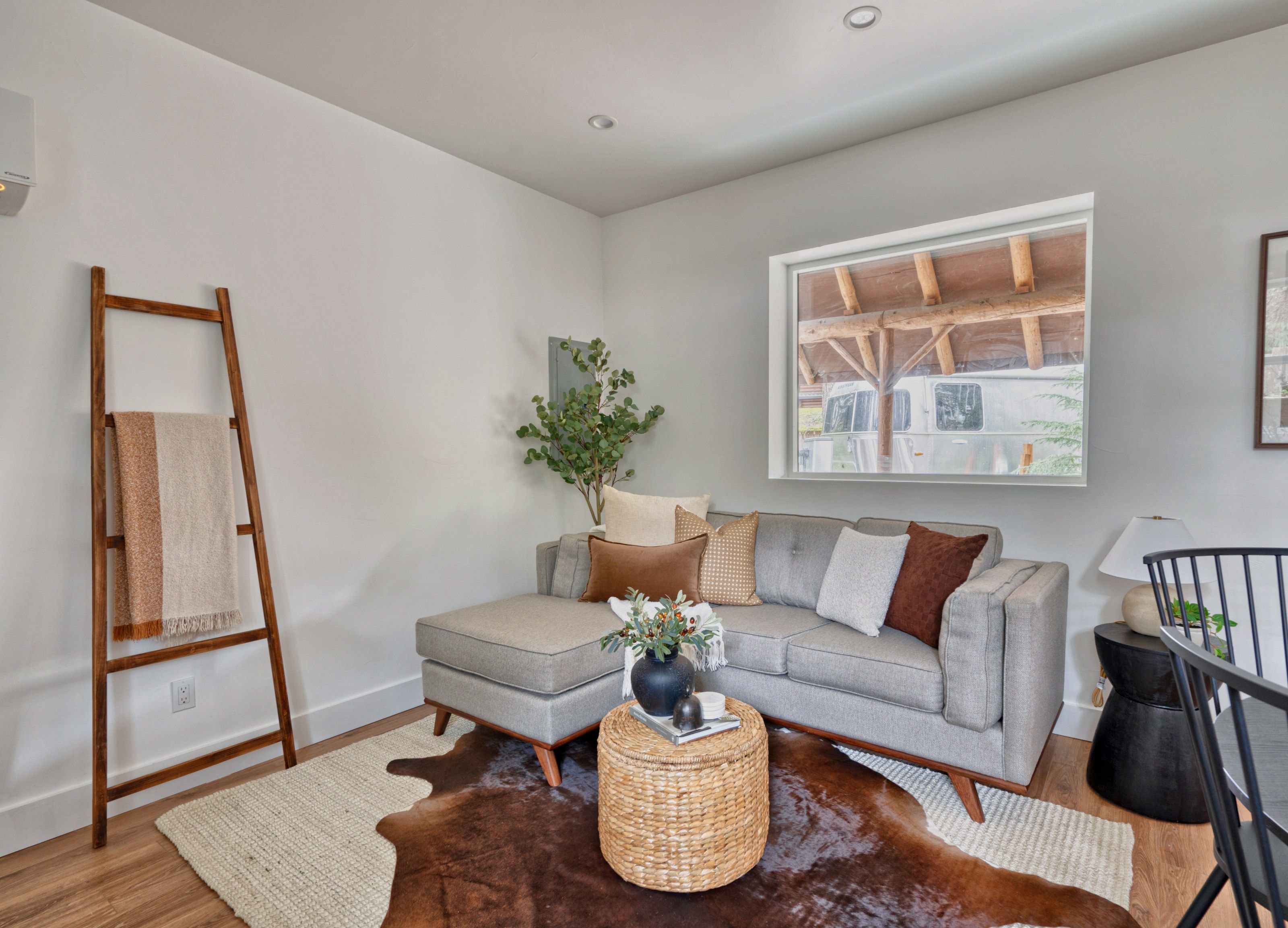 A modern living room with a grey sofa, a wooden ladder, a cowhide rug, and a window with a view of a building.