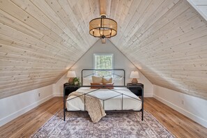 Een slaapkamer met een schuine kap, een bed met een geometrisch hoofdeinde, twee nachtkastjes met lampen, een kamerkleed met patroon, en een raam met uitzicht op bomen.