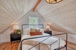 Une chambre avec un plafond en pente, un lit avec un cadre en métal, deux tables de chevet avec des lampes et une fenêtre donnant sur les arbres.