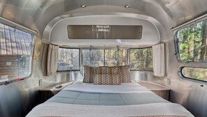 Ein kompakter, gut beleuchteter Innenraum mit einem Bett, einem kleinen Tisch und einem Fenster mit Blick auf einen Wald.