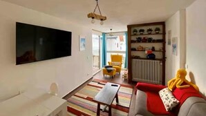 Ein Wohnzimmer mit Flachbildfernseher, einer Couch, einem Couchtisch, einem gelben Sessel, einem Bücherregal und einem Fenster mit Vorhängen.