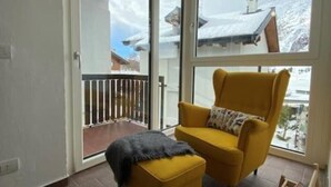 Ein Zimmer mit einem gelben Sessel, einem kleinen Hocker und einem Holztischchen. Ein großes Fenster bietet Blick auf schneebedeckte Häuser.