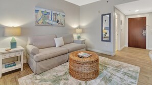 A living room with a beige sofa, two side tables with lamps, a wicker ottoman, a painting of surfboards, and a wooden door.