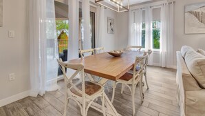 A dining area with a wooden table and white chairs, a chandelier, and a view through large windows.