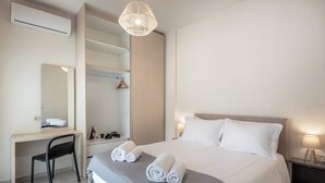 A hotel room with a bed, a desk, a chair, a wardrobe, a hanging light fixture, and a bedside table with a lamp. - Harma City Living - Street View Family Apartment (Chania)
