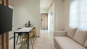 A modern living room with a sofa, a dining table with chairs, a flat-screen TV, and a kitchen area with a coffee maker. - Harma City Living - Street View Family Apartment (Chania)