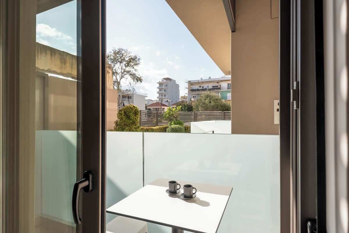 Een balkon met een glazen deur, een kleine tafel met twee kopjes en uitzicht op gebouwen en bomen.