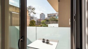Een balkon met een glazen deur, een kleine tafel met twee kopjes en uitzicht op gebouwen en bomen.