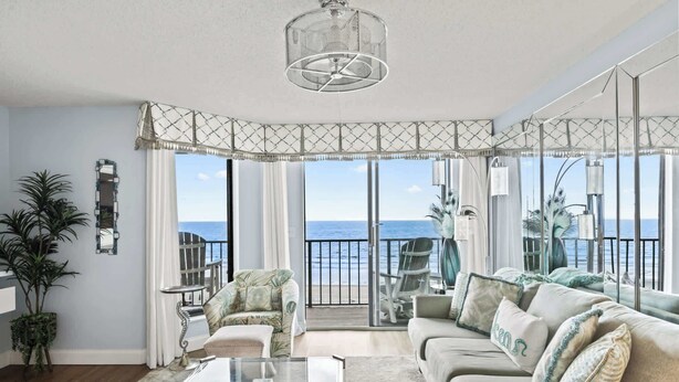A spacious living room with a large window overlooking the sea, a ceiling fan, and a glass coffee table. - Beach Club I Unit 5F - Oceanfront! (North Myrtle Beach)