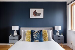 Een moderne slaapkamer met een donkerblauwe wand, een groot bed met een grijze hoofdbord en twee nachtkastjes met lampen en decoratieve kussens.