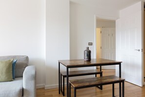 Een kamer met een grijze bank, een houten tafel met een bankje, en een witte deur op de achtergrond.