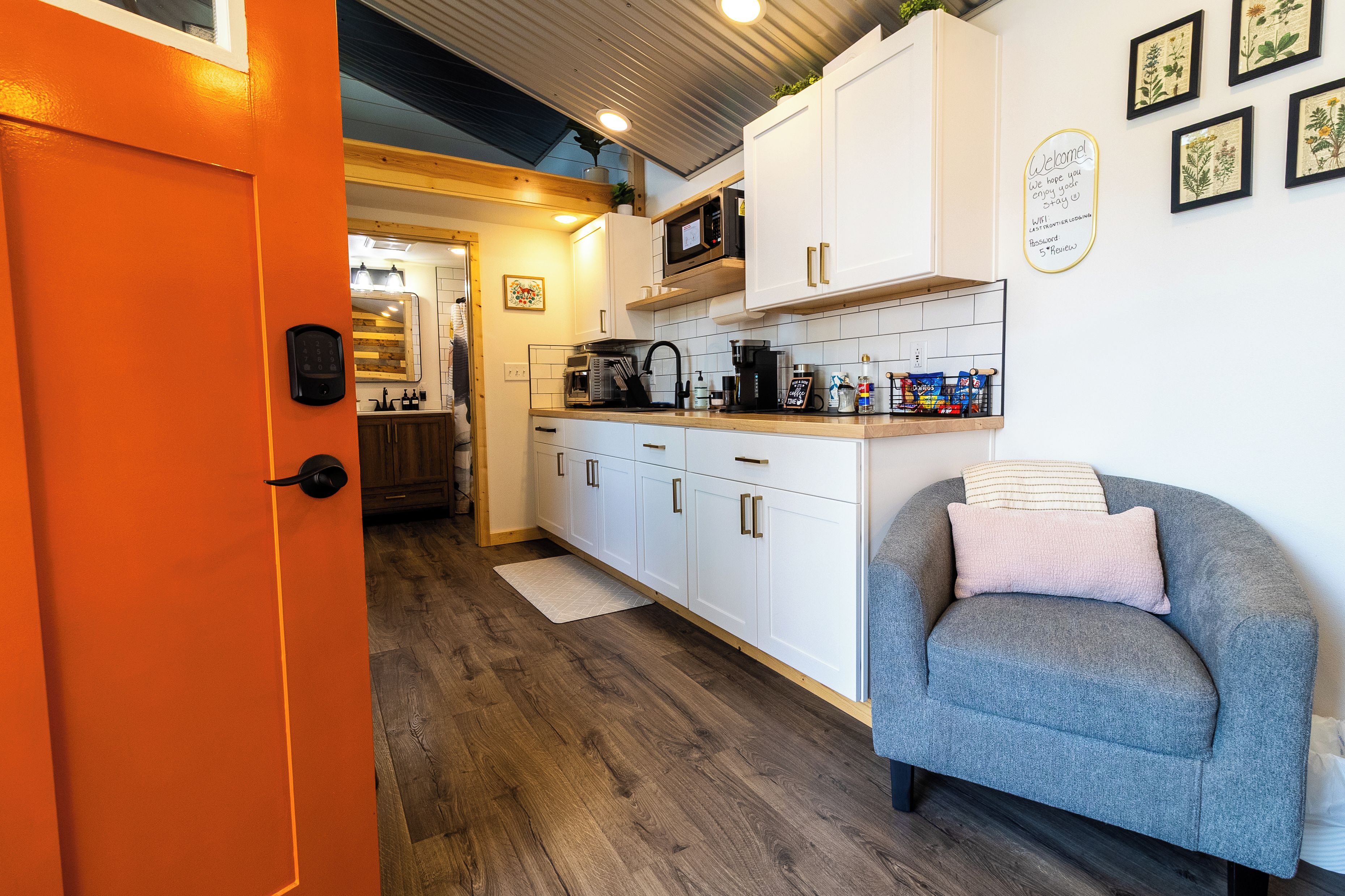 A compact kitchen with white cabinets, a microwave, and a coffee maker. There is a grey sofa with a pillow and a small rug on the floor.
