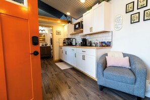 A compact kitchen with white cabinets, a microwave, and a coffee maker. There is a grey sofa with a pillow and a small rug on the floor. - Orange Poppy - Seward Alaska Modern Forest Tiny Home Retreat Near Kenai Fjords (Seward)