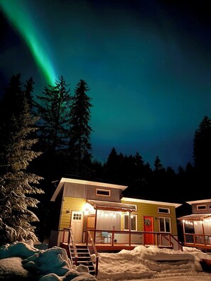 A two-story building with a snow-covered ground, a staircase leading to the entrance, and a view of the night sky with the aurora borealis. - Orange Poppy - Seward Alaska Modern Forest Tiny Home Retreat Near Kenai Fjords (Seward)