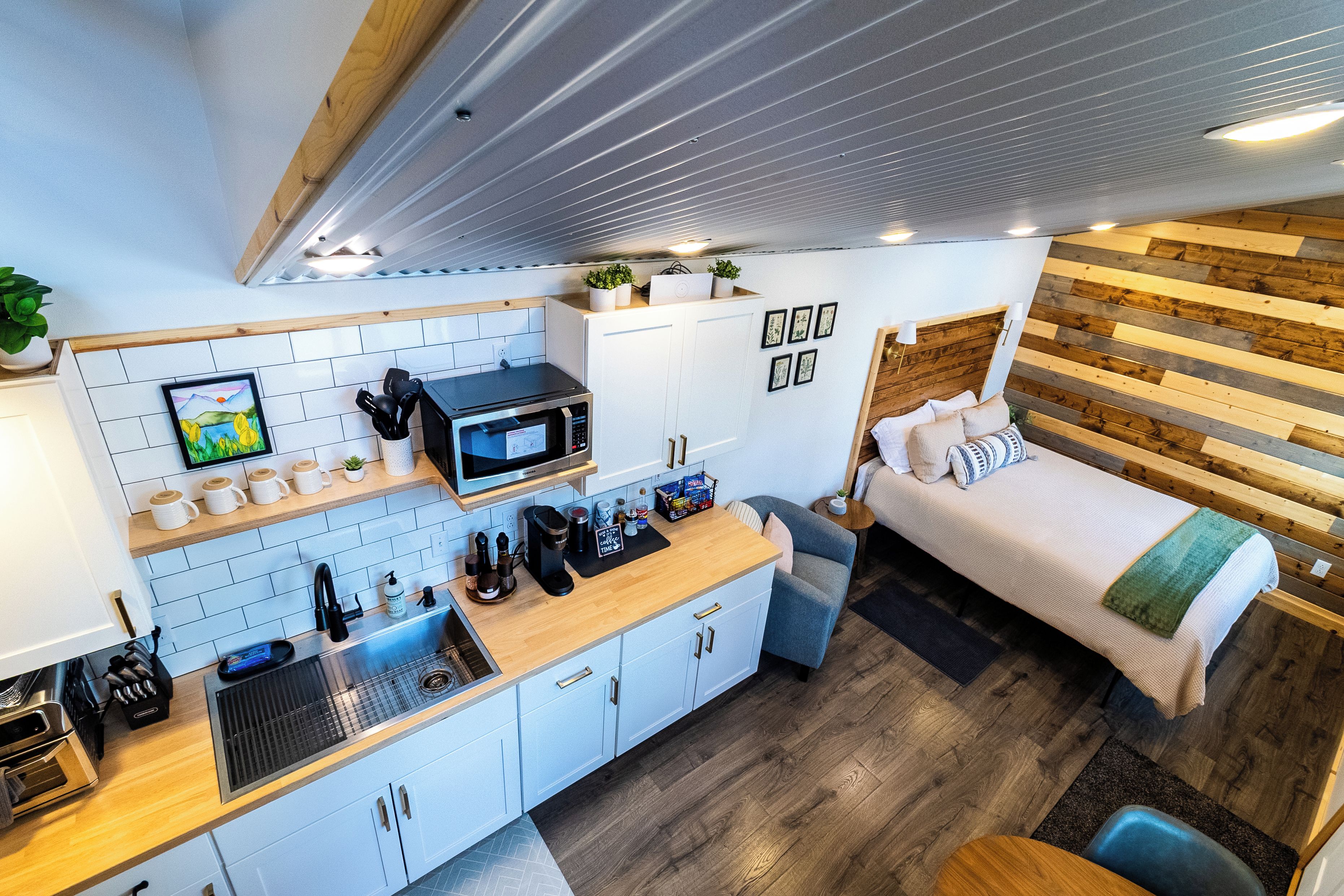 Een compacte, moderne kitchenette met een magnetron, koffieapparaat en diverse apparaten. Er is een klein zithoekje met een bank en een bed met een houten hoofdbord.
