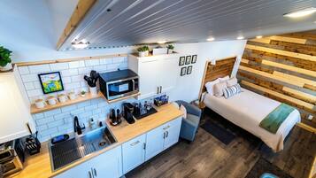Een compacte, moderne kitchenette met een magnetron, koffieapparaat en diverse apparaten. Er is een klein zithoekje met een bank en een bed met een houten hoofdbord.